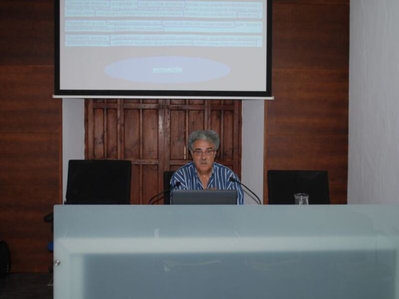 Persona impartiendo una conferencia o presentación en un salón con pantalla de proyección en el fondo.