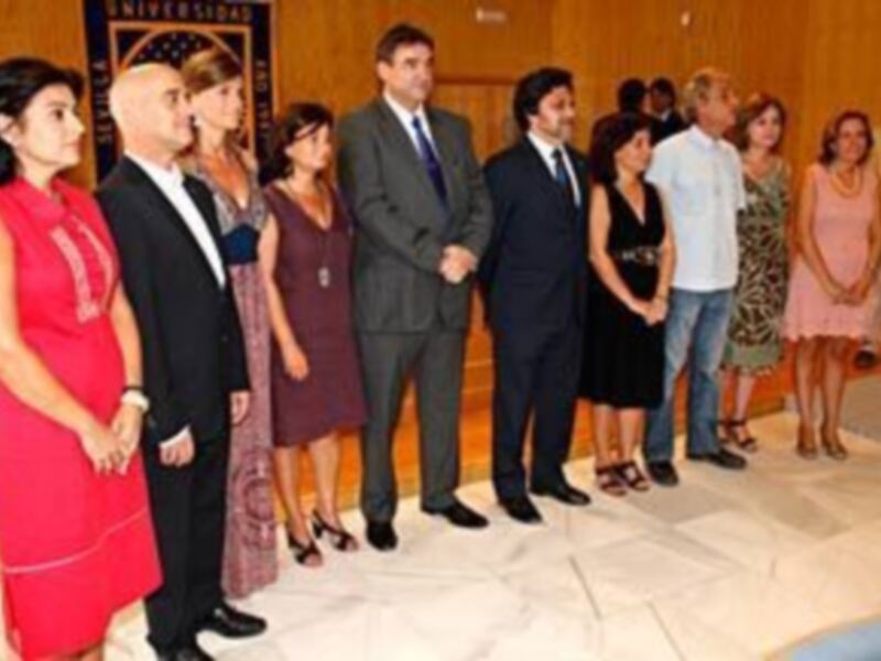 Un grupo de personas forman una fila en un evento formal, vestidas con trajes y vestidos de colores variados, posando para una foto en una sala decorada.