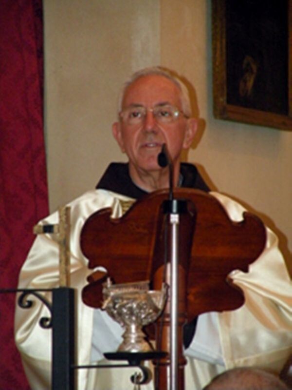 Elderly man in religious attire, possibly a priest, standing at a lectern with a microphone and a chalice in front of him.