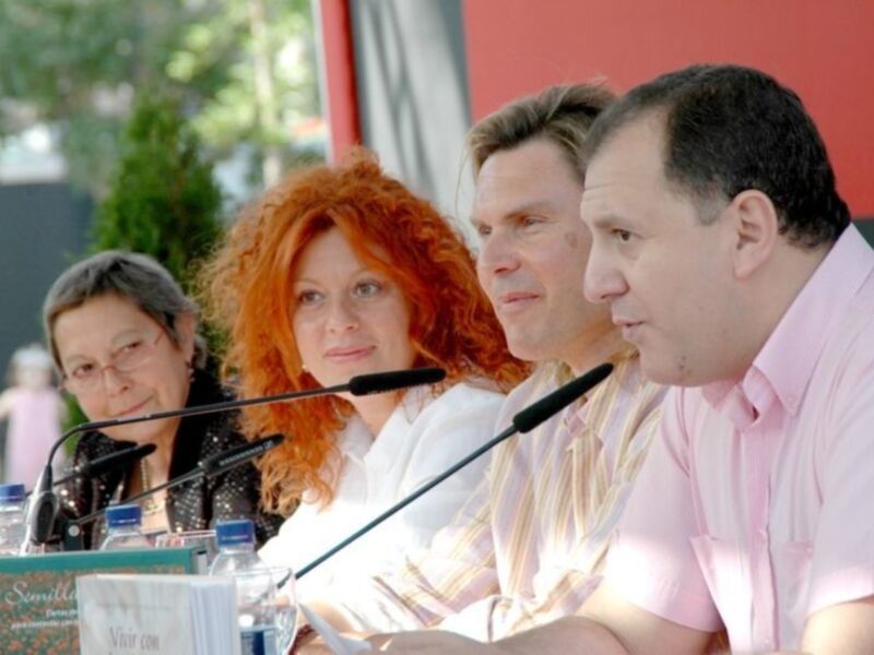 Una escena formal con cuatro personas sentadas en una mesa, cada uno frente a un micrófono. La imagen parece ser de una conferencia o evento público, con el fondo rojo y la presencia de un microtelón.