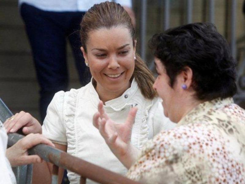Una mujer sonríe mientras habla con otra persona en una escalera. La escena parece ser en un entorno urbano con estructuras de hormigón en el fondo.