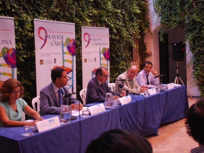 Una conferencia de prensa en un evento llamado 'Cursos de Verano 2011', con personas sentadas en una mesa con microfonos y carteles promocionales en el fondo.