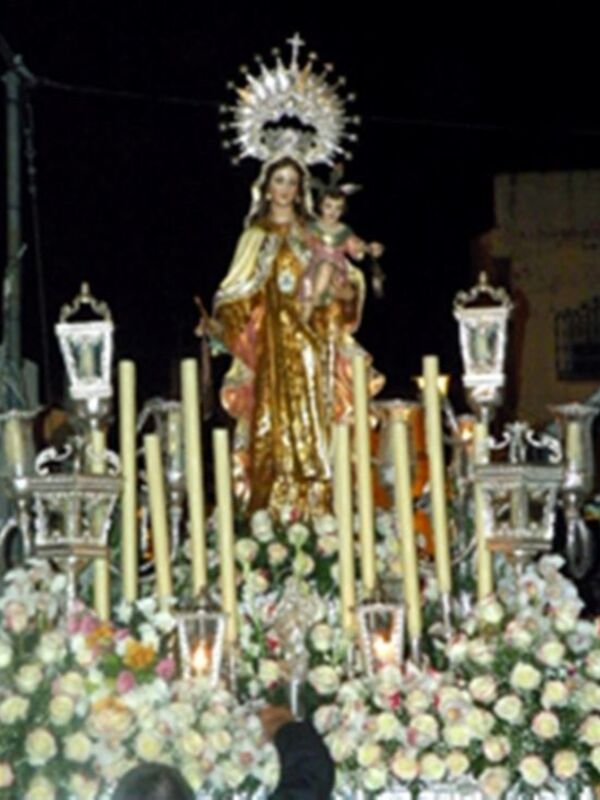 Estatua de la Virgen con el Niño en una procesión nocturna, rodeada de flores y luces.