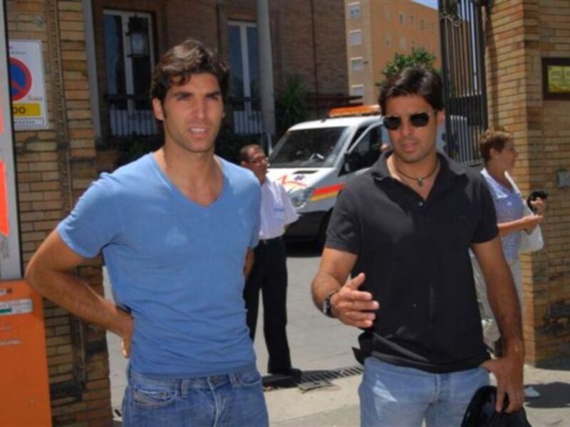 Dos hombres caminando por una calle urbana, uno con camiseta azul y el otro con polo negro, en un entorno urbano con edificios y vehículos al fondo.