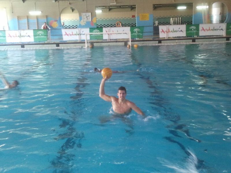 Un hombre sostiene una pelota de waterpolo en una piscina interior, con otros nadadores en el fondo y un muro decorado con carteles de Andalucía.