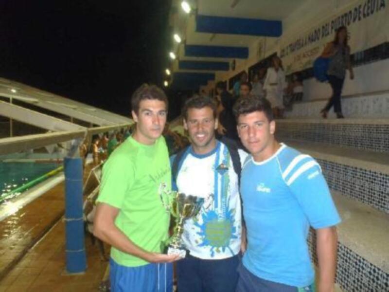 Tres personas posan con un trofeo en un evento deportivo nocturno en el Puerto de Ceuta.