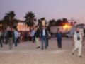 Una multitud camina por una zona de playa, con palmeras y edificios en el fondo. La imagen muestra a varias personas de diferentes edades, incluyendo hombres y mujeres, algunas con mochilas y otros con bolsos. La iluminación sugiere que es tarde o temprano en la noche, y el ambiente es casual.