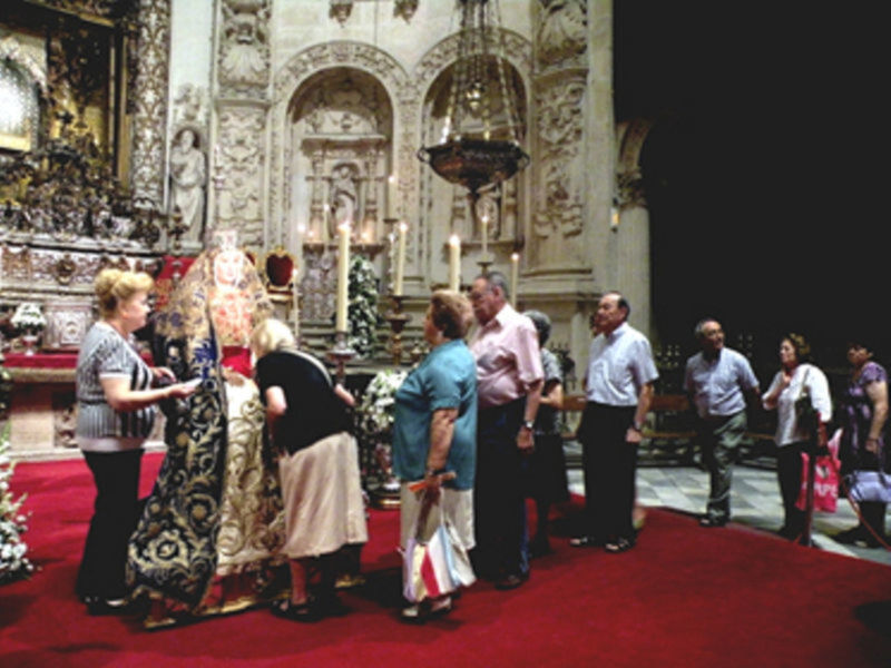 Gente en una iglesia, con una figura religiosa en el fondo y una alfombra roja en el suelo. La iglesia tiene detalles artísticos y una arquitectura elaborada.