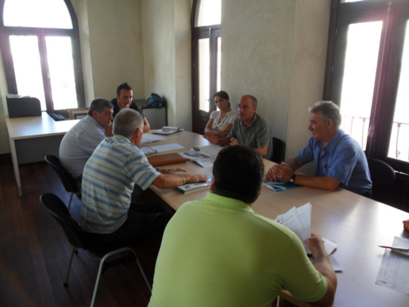 Un grupo de personas reunidas en una sala con ventanales, discutiendo y trabajando en documentos en un escritorio. La atmósfera es profesional y concentrada.