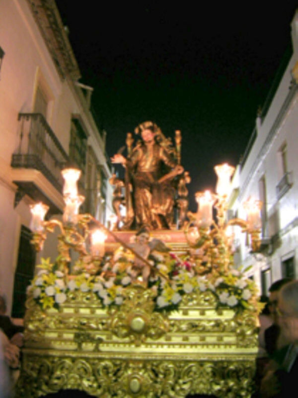 Procesión nocturna con estatua dorada en un carro ornado con flores y candelabros iluminados.