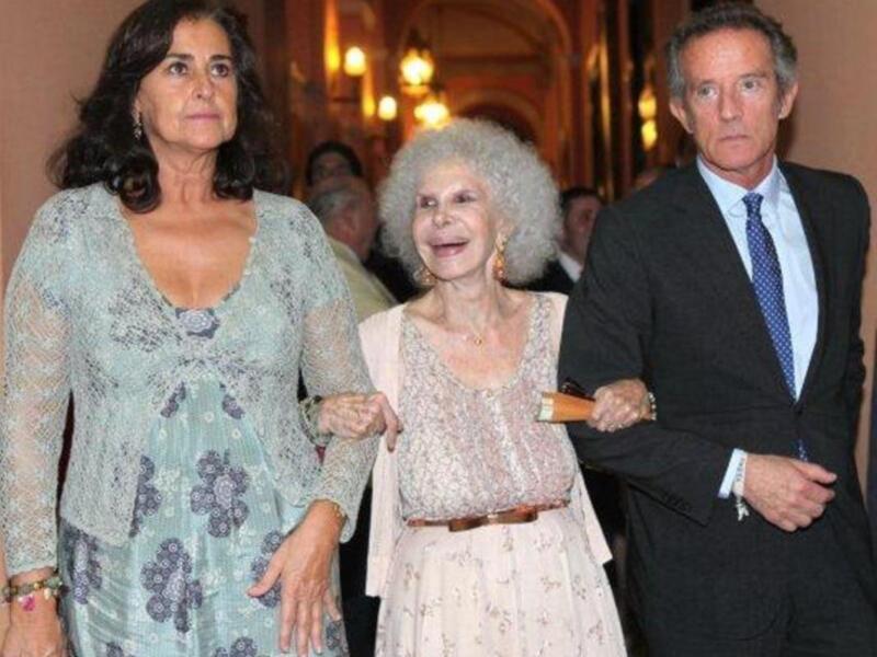 Tres personas caminando juntas en un evento formal, vestidas elegantes, en un ambiente iluminado con lámparas.