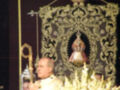 Una persona vestida con un hábito sacerdotal, posiblemente en una ceremonia religiosa, frente a un altar con flores y una corona.