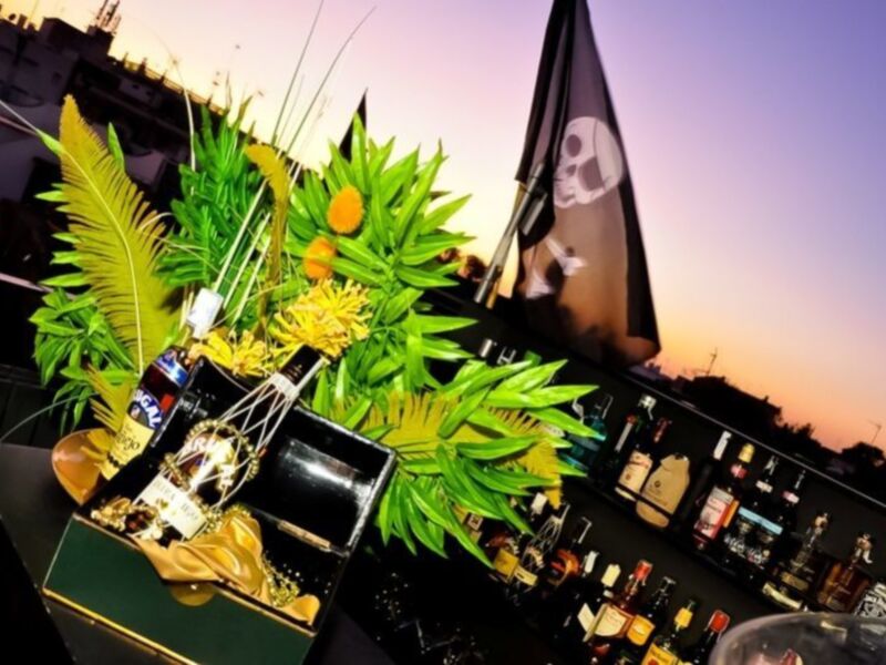 Barra de botellas de cerveza y licores con bandera pirata en el fondo, iluminación de atardecer en el cielo.
