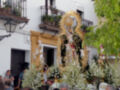 Una procesión religiosa en un entorno urbano, con una figura de la Virgen María en el centro. La procesión está acompañada por flores y decoraciones, y se realiza frente a un edificio con balcones. La atmósfera es festiva, típica de celebraciones religiosas en España.