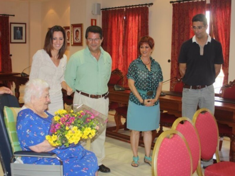 Una anciana en silla de ruedas recibe una floración de flores de manos de una mujer mientras un hombre y un joven observan la escena.