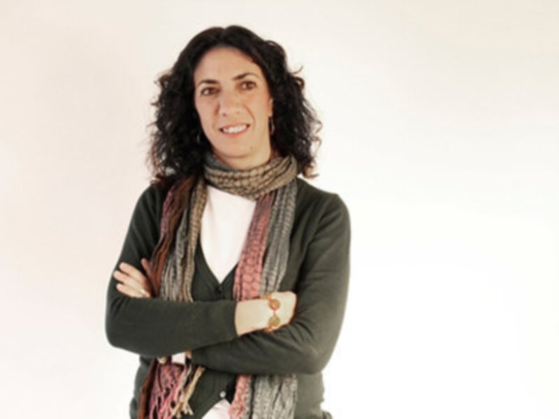 Retrato de una mujer sonriente con brazos cruzados, vistiendo una bufanda y una chaqueta verde, contra un fondo blanco.
