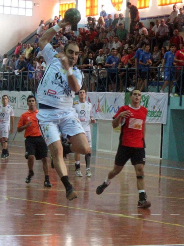 Jugador de balonmano en acción durante un partido, lanzando el balón mientras el público observa en un estadio cubierto.