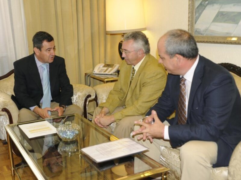 Tres hombres en trajes sentados en un sofá, discutiendo en una sala formal con una mesa de cristal y documentos.