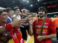 Un jugador de baloncesto español celebra con su equipo y seguidores en un estadio lleno. El jugador lleva una camiseta roja con el nombre "España" y un cinturón verde. La camiseta también tiene la palabra "Campeones" y el año 2015. Los seguidores son jóvenes con camisetas rojas y una bandera de España. La arena está iluminada y el público es multitudinario.