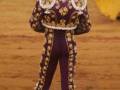 Un torero en un traje tradicional de color morado con detalles dorados, observando la arena mientras sostiene una bandera rosa y amarilla.