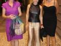 Tres mujeres posan juntas en un evento formal, dos de ellas con vestidos largos y una con un vestido negro. La imagen muestra detalles como joyas, bolsos y zapatos de tacón.
