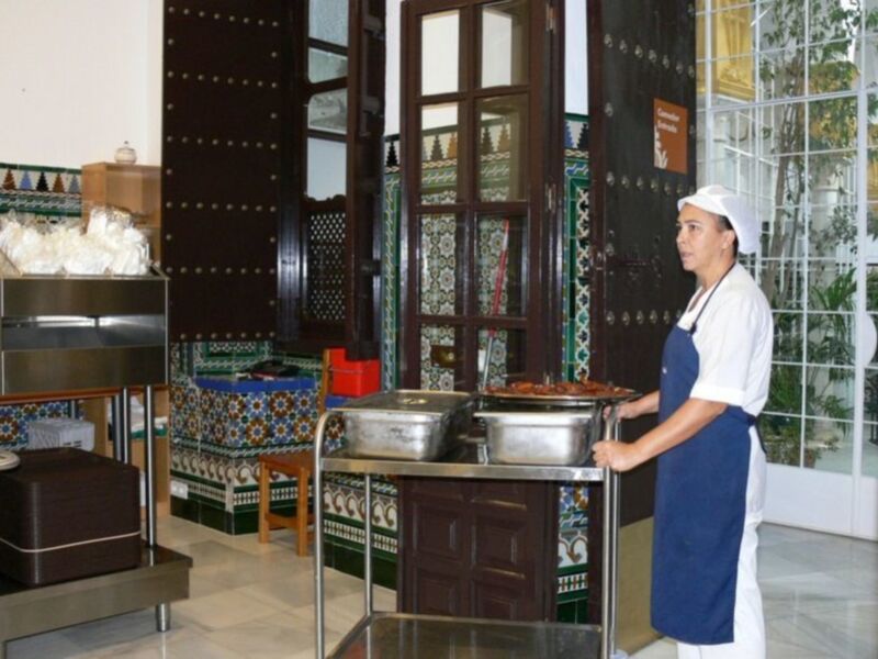 Trabajador de restaurante con uniforme blanco y azul, transportando bandejas de comida en un ambiente decorado con azulejos tradicionales.
