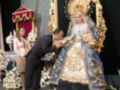 Una imagen de una figura religiosa vestida con un manto azul y dorado, posiblemente una Virgen, en un altar adornado con flores blancas y un hombre y una mujer observando la escena.