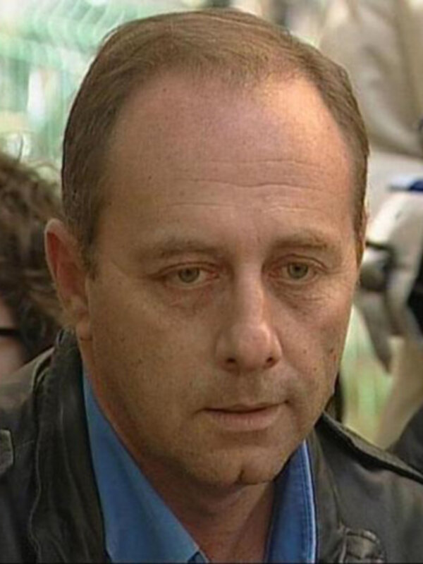 Un hombre con una chaqueta negra y una camisa azul, posiblemente en una situación de entrevista o conferencia de prensa.