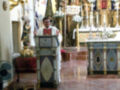 Una imagen de un altar en una iglesia con un sacerdote vestido con un hábito blanco y rojo, frente a una pila bautismal. El altar está decorado con detalles dorados y azules, y hay flores en el fondo.