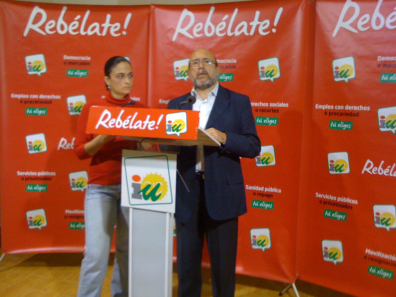 Un hombre y una mujer hablando en un evento político con pancartas rojas y el lema 'Rebélate!'.