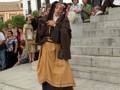 Una mujer vestida con una túnica marrón y falda beige, posiblemente en un evento histórico o cultural. Detrás de ella, se observan personas y una estructura blanca con detalles arquitectónicos. La escena se desarrolla en un entorno urbano, con una calle adoquinada y escalones de piedra.