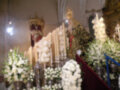 Imagen de una iglesia con altares decorados con flores blancas y velas. En el centro, una imagen de Cristo rodeada por adornos dorados y flores. A la izquierda, una imagen de San Juan Evangelista con vestimenta roja y capa blanca. A la derecha, un altar con flores y velas blancas. La atmósfera es solemnemente festiva, probablemente durante una celebración religiosa o procesión.
