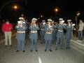 Un grupo de músicos militares en uniformes azules y sombreros blancos toca instrumentos musicales al aire libre. La noche está oscura, y las luces de la ciudad se pueden ver en el fondo.