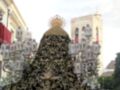 Imagen de una procesión religiosa con un paso de Cristo en la Plaza de España, Sevilla. El paso está decorado con detalles dorados y una corona en la parte superior. La torre de la catedral se puede ver al fondo, rodeada por personas observando la procesión.