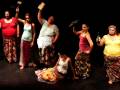 Una escena teatral con cinco actrices vestidas con diversos estilos de saris y pantalones, sosteniendo objetos como un martillo, una botella y otros. La actriz en el centro está sentada con una expresión de dolor o fatiga, mientras las demás actrices muestran gestos de protesta o frustración.
