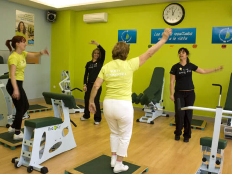 En un gimnasio, varias personas participan en una clase de ejercicios. La pared está decorada con carteles informativos y un reloj. La escena transmite una atmósfera de actividad física y motivación.