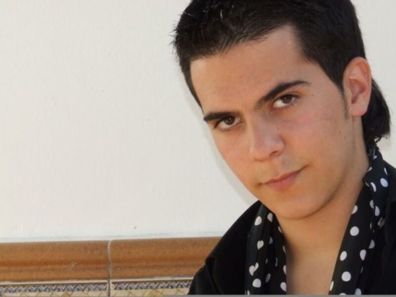 Un hombre joven con una camisa negra y una bufanda de lunares blancos, posando frente a una pared blanca y un marco de fotos.