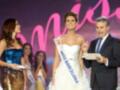 A una mujer con un vestido blanco y un cinturón dorado, sosteniendo un trofeo con el texto "Miss Barcelona". A su lado, hay una mujer vestida de azul brillante y otra con un vestido blanco. Un hombre en traje negro sostiene una hoja de papel y parece estar anunciando algo. El fondo es rosado con letras que parecen "Miss".