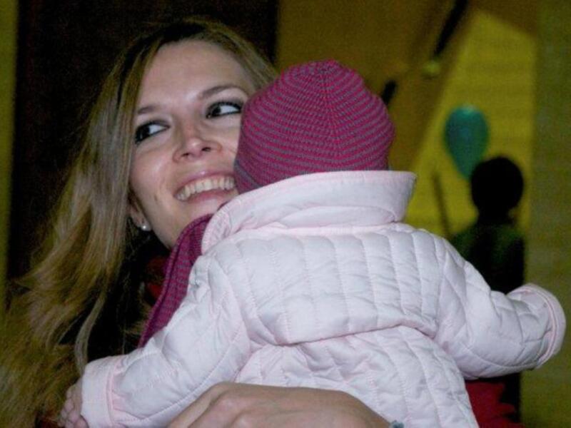 Una mujer sonríe mientras abraza a un bebé vestido con un suéter rosa y una gorrita tejida de color rosa y blanco.