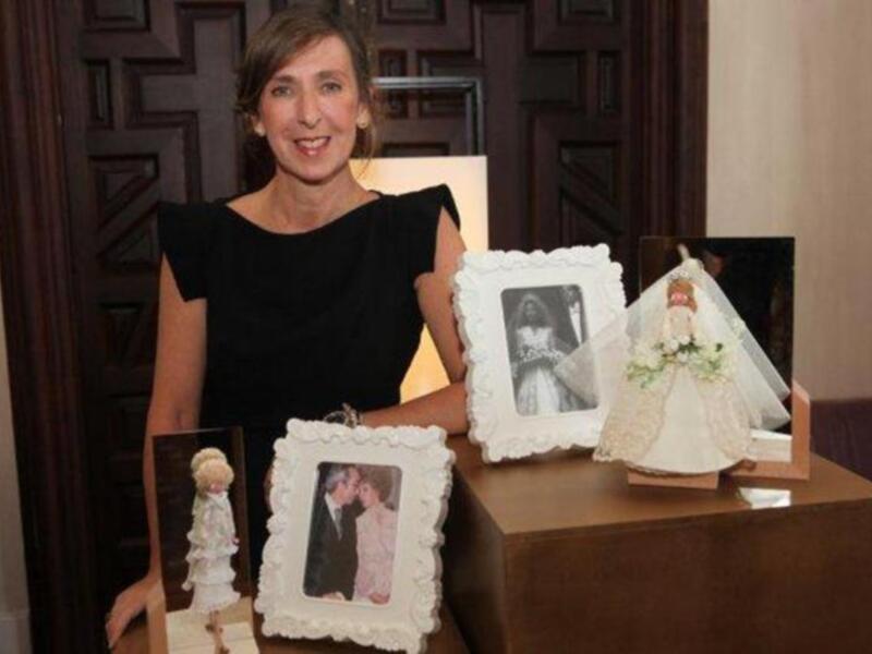 Mujer con vestido negro posando junto a una mesa con fotos y una muñeca vestida de blanco.