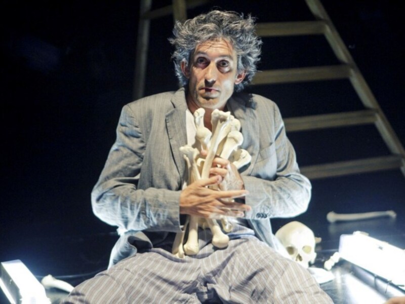 Actuador en escena sosteniendo huesos humanos, con un entorno teatral oscuro y un cráneo en el fondo.