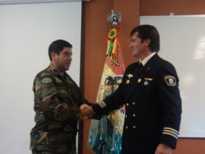 Dos militares se saludan en una ceremonia formal, frente a una bandera y un tablero blanco.