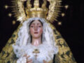 Imagen de una Virgen María vestida con un manto dorado y una corona de estrellas, sosteniendo un cáliz. La escena se desarrolla en una iglesia con una vela encendida a su lado.