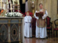 Imagen de una ceremonia religiosa en un altar con flores blancas y rojas, un sacerdote vestido de blanco y una niña en vestido blanco.
