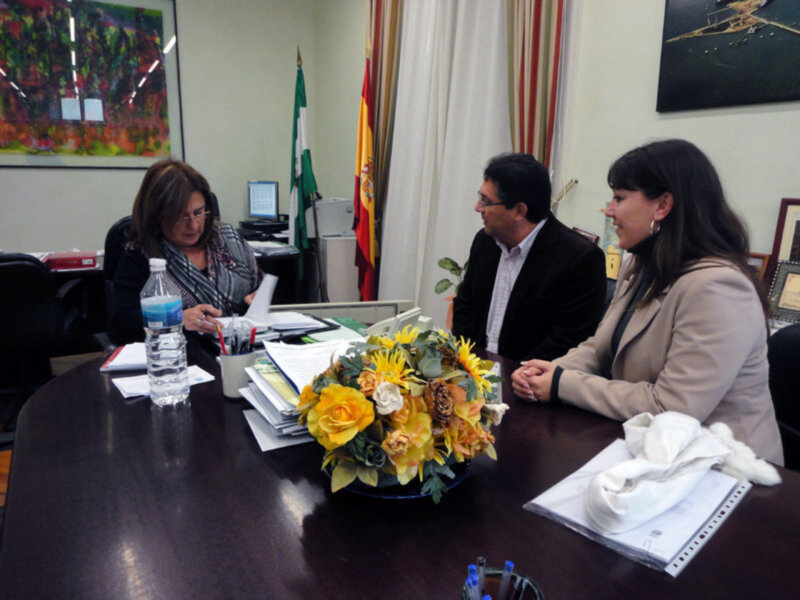 Dos personas en una reunión, con una bandera española en el fondo y un jarrón de flores en el centro de la mesa.