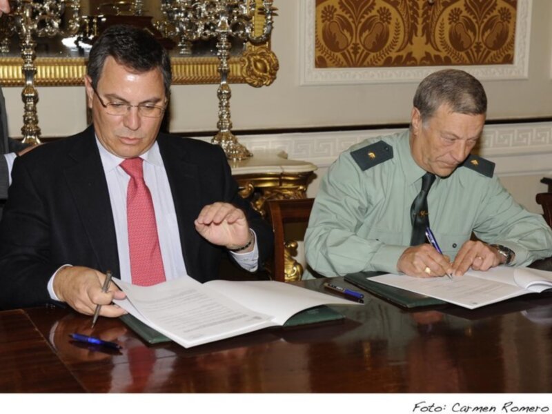 Dos hombres firmando documentos en una sala formal con una mesa de madera y una decoración de estilo clásico.