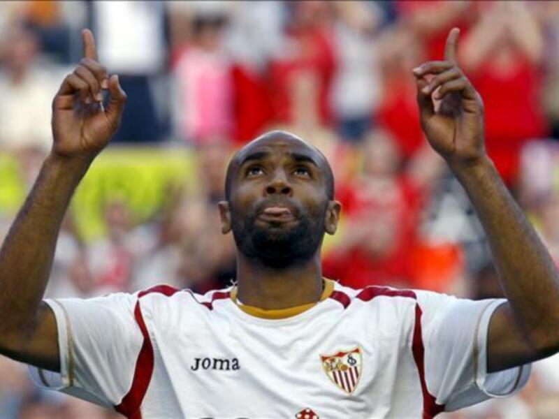 Un jugador de fútbol celebra con los brazos levantados, luciendo un uniforme blanco y rojo con el escudo del Sevilla FC en el pecho.