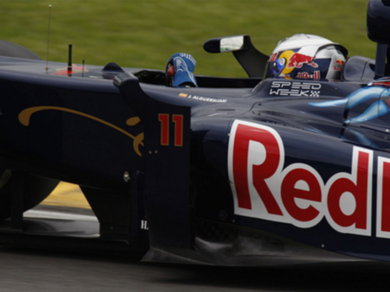 Fórmula 1 carrera en un circuito, con un piloto en el cockpit del coche. El coche tiene una bandera de Reino Unido y la palabra 'RedBull' en la parte trasera.