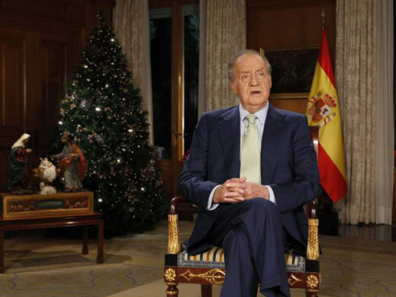 El Rey Juan Carlos en una toma de Navidad en el Palacio Real de Madrid, con una bandera española y un árbol de Navidad en el fondo.