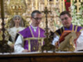 Dos sacerdotes vestidos con vestiduras religiosas de color púrpura y dorado, uno leyendo un libro mientras el otro mira hacia la cámara. En el fondo, una estatua de una figura vestida con un manto blanco y tocado con un halo dorado, junto a elementos decorativos como velas y flores.